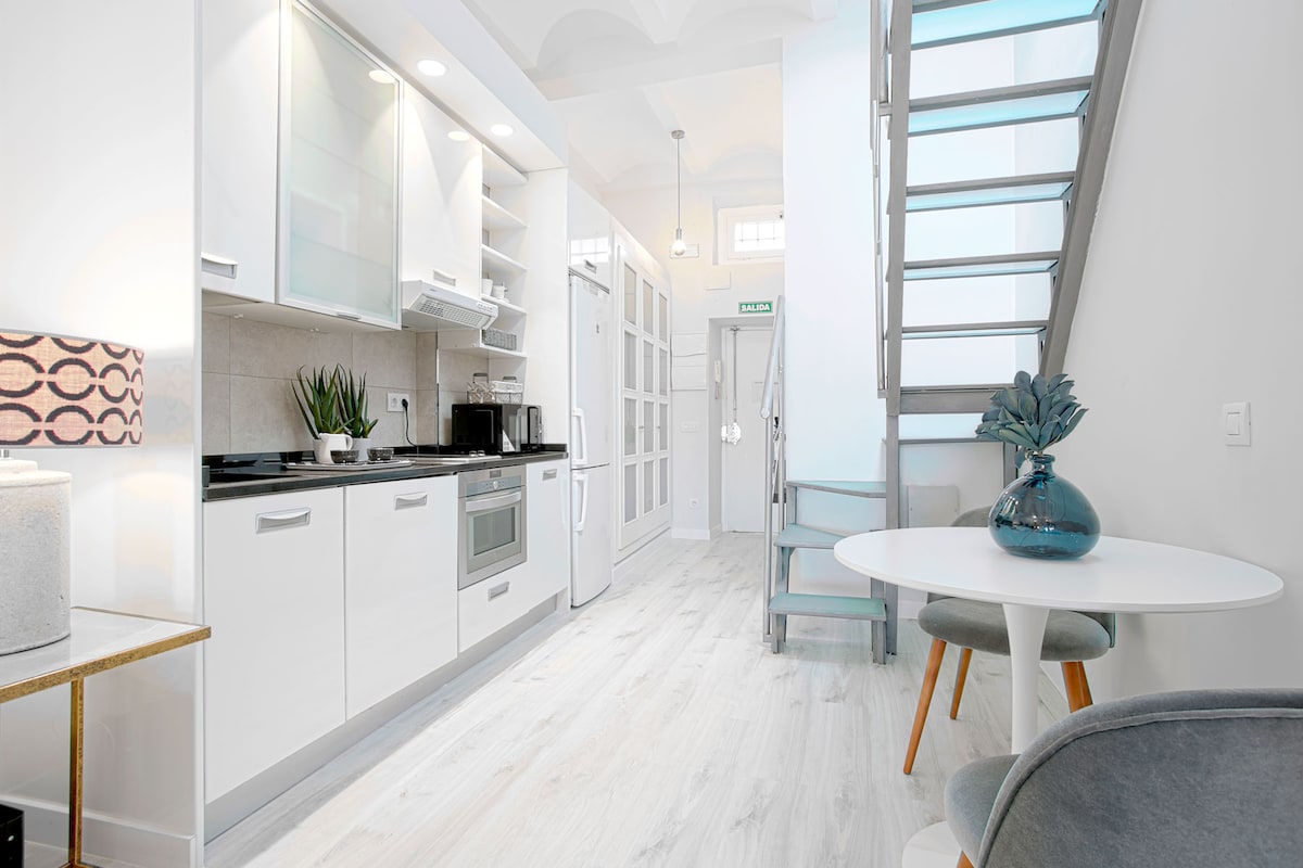 The kitchen area features modern appliances and sleek cabinetry, complemented by a dining table with two chairs. Natural light floods the space through large windows, and a spiral staircase provides access to the upper floor. Contemporary décor enhances the stylish ambiance.