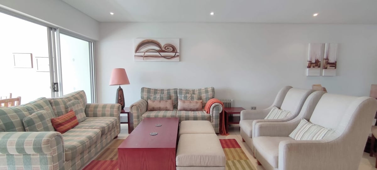 A bright living area features a combination of plaid and neutral-toned sofas, along with two light-colored armchairs. A wooden coffee table sits at the center, complemented by a colorful area rug. Light enters through large sliding glass doors, enhancing the spacious atmosphere.