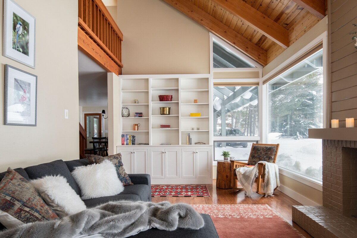 A cozy living area features a dark couch adorned with soft throw pillows and a plush blanket. A white built-in shelf holds decorative items, while large windows allow natural light to fill the space and provide views of the snowy outdoors. Warm wood accents enhance the atmosphere.