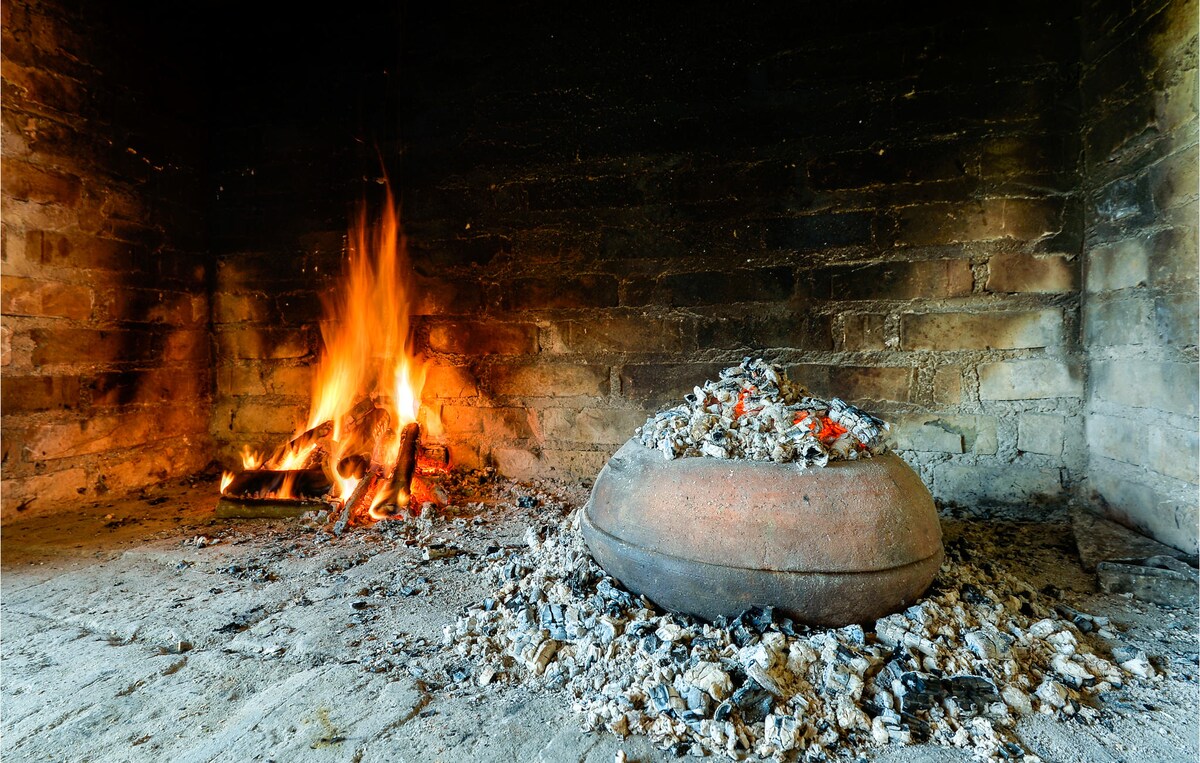 A traditional fireplace is seen, with flames rising from logs set in a stone hearth. A round clay pot rests on a bed of ashes, surrounded by a rustic earthen floor and brick walls, suggesting an ambiance conducive to warmth and rustic cooking.