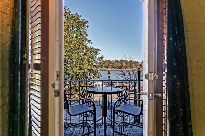 Double doors open to a serene balcony view overlooking the river. A small round table and two chairs are positioned on the balcony, inviting relaxation. Lush greenery and a clear blue sky create a peaceful backdrop for enjoying outdoor moments.