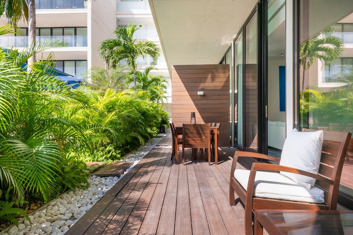 A private deck is bordered by lush greenery, featuring a wooden table and chairs for outdoor dining. Decorative white stones line the edge, and a comfortable chair with a cushion is positioned nearby. Floor-to-ceiling glass doors provide a seamless connection to the interior.