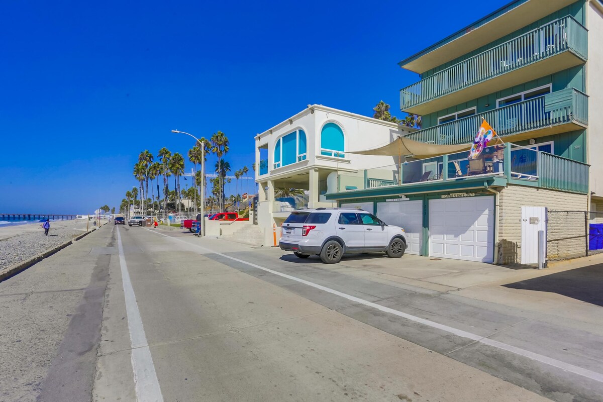 The image captures the exterior of a three-story building located near the beach. A wide street lined with palm trees runs alongside. The building features balconies and garages, with a white SUV parked in front. Bright blue skies create a sunny atmosphere.
