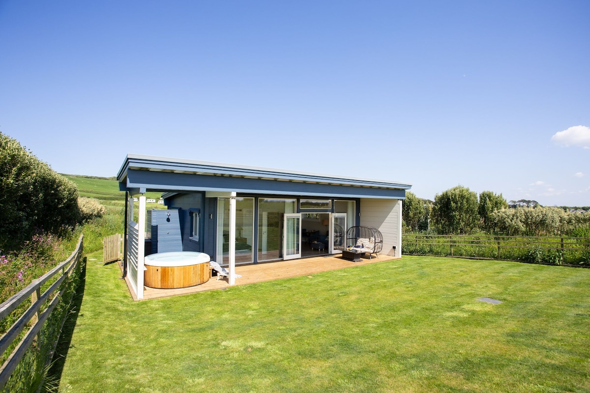 A modern lodge features a spacious decked patio, with a private hot tub visible beside the exterior. The surrounding lawn is lush and well-kept, offering a serene view of nearby greenery and blue skies.