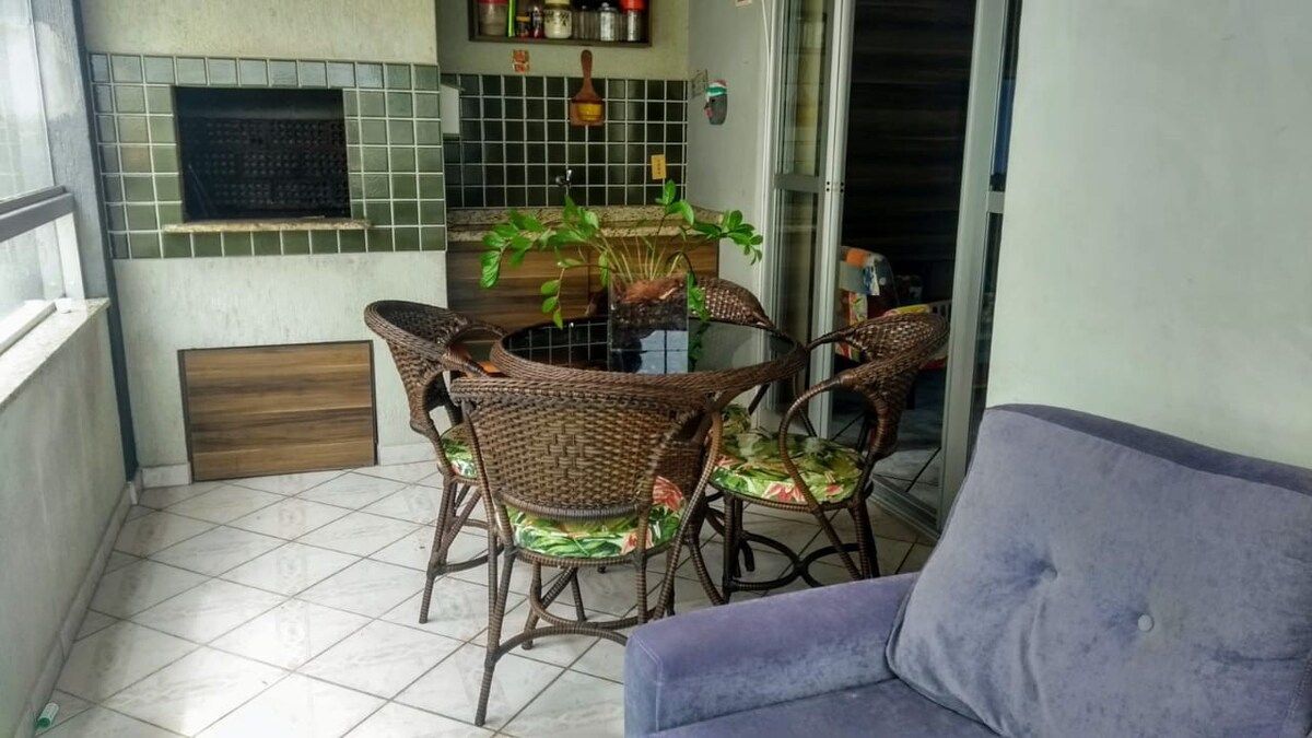 An outdoor balcony features a charcoal grill and a round dining table surrounded by four woven chairs with floral cushions. A plush blue sofa is positioned nearby, while light-colored tiles and large windows create an airy feel in the space.