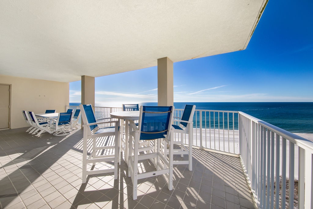 A spacious balcony is furnished with white and blue outdoor furniture, including tables and chairs. The ocean view stretches towards the horizon under a clear blue sky, inviting guests to enjoy the coastal atmosphere.