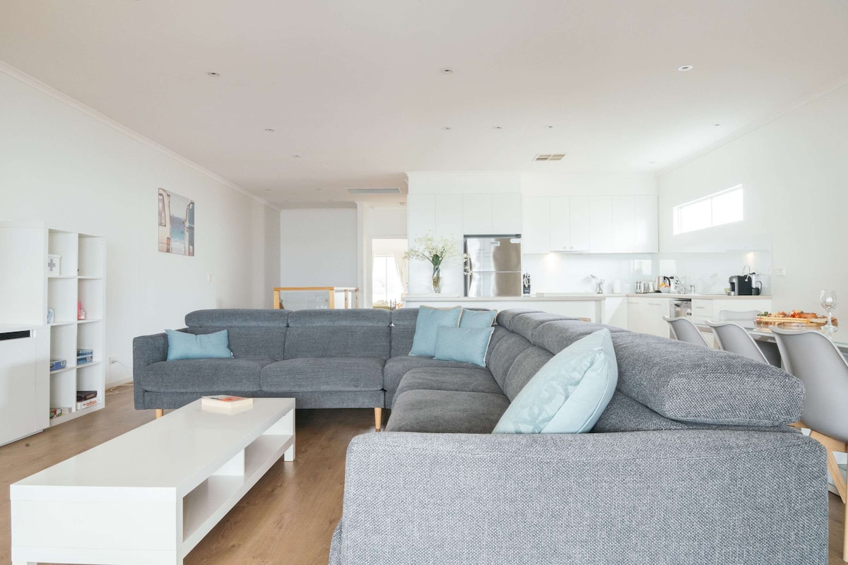 A spacious living area is presented, featuring a large gray sectional sofa with light blue accents. A white coffee table is centered in the space. The open-plan layout connects to a bright kitchen, visible in the background, with white cabinetry and modern appliances.