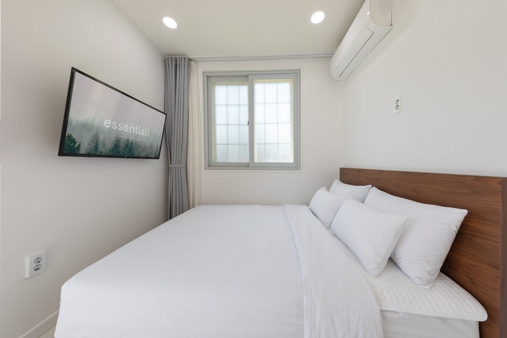 A light-filled room features a spacious bed, dressed in crisp white linens and multiple pillows. A flat-screen television is mounted on the wall opposite the bed. A large window allows natural light to brighten the space, while sheer curtains provide privacy.