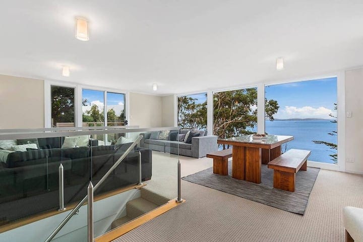 An open living area is showcased, featuring large windows that offer expansive water views. A wooden dining table with bench seating is centrally positioned, complemented by a comfortable grey sofa. Soft carpeting covers the floor, creating a welcoming atmosphere.