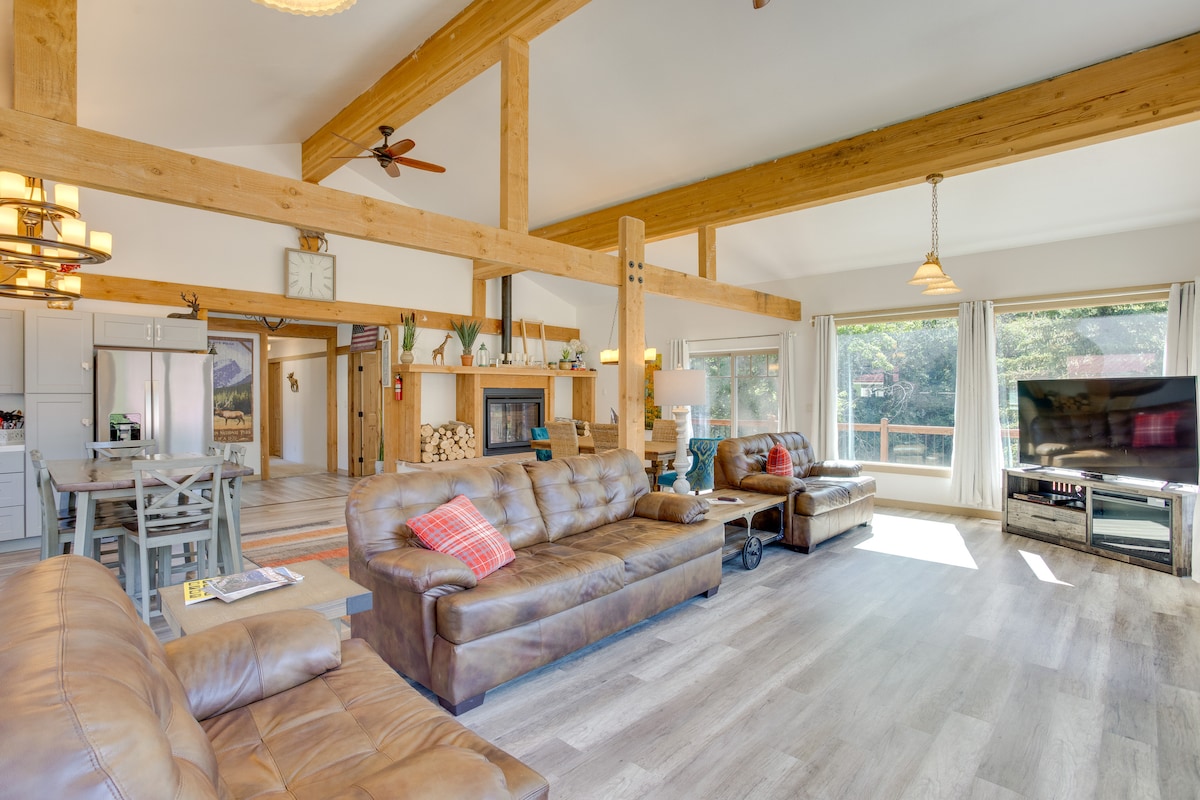 A spacious living area showcases wooden beams and large windows allowing natural light to fill the space. Comfortable leather sofas are arranged around a coffee table, while a fireplace is framed by a decorative wall. A dining area and kitchen are visible in the background.