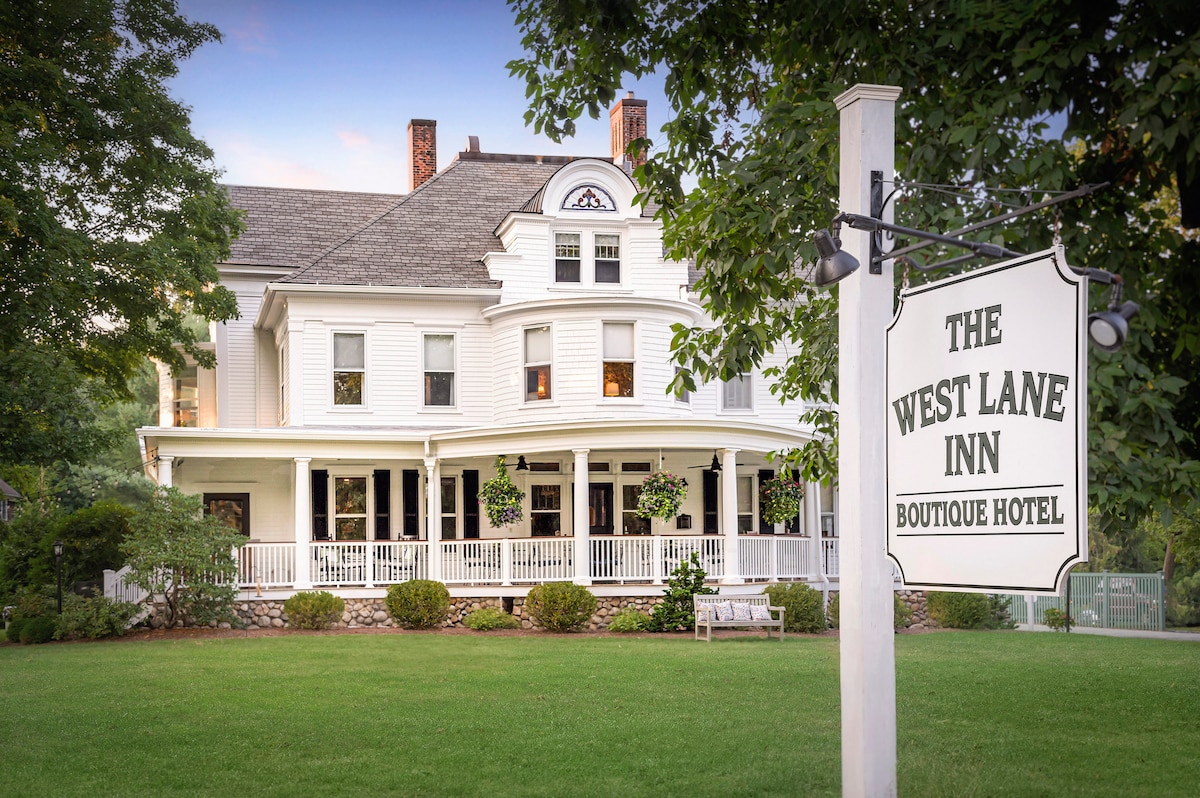 An elegant white inn is showcased surrounded by a lush green lawn, framed by trees and flower beds. The iconic sign displays 'The West Lane Inn Boutique Hotel.' A spacious porch with white railings is visible, inviting guests to relax in a serene outdoor setting.