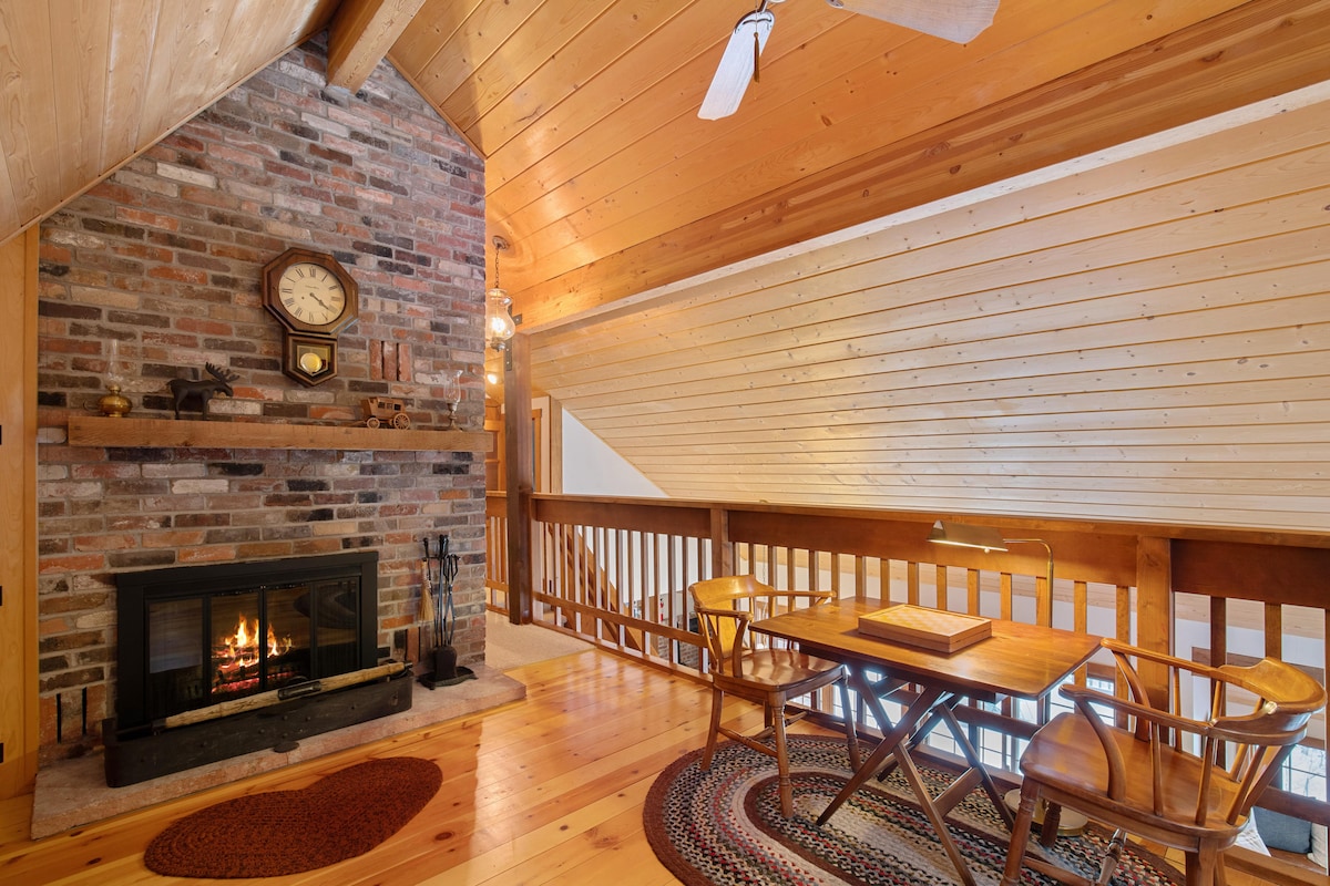 A cozy loft area showcases a wood-burning fireplace set against a brick wall, complemented by a rustic clock. Natural wood paneling and flooring create a warm ambiance. A small table with two chairs sits nearby, inviting relaxation while enjoying the views of the surrounding space.