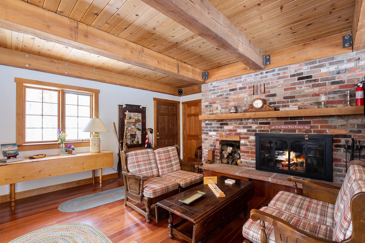 A welcoming living room features wooden beams and a brick fireplace, enhancing the rustic charm. Comfortable seating is provided by plaid chairs and a wooden coffee table. Natural light enters through the windows, illuminating the warm wood tones throughout the space.