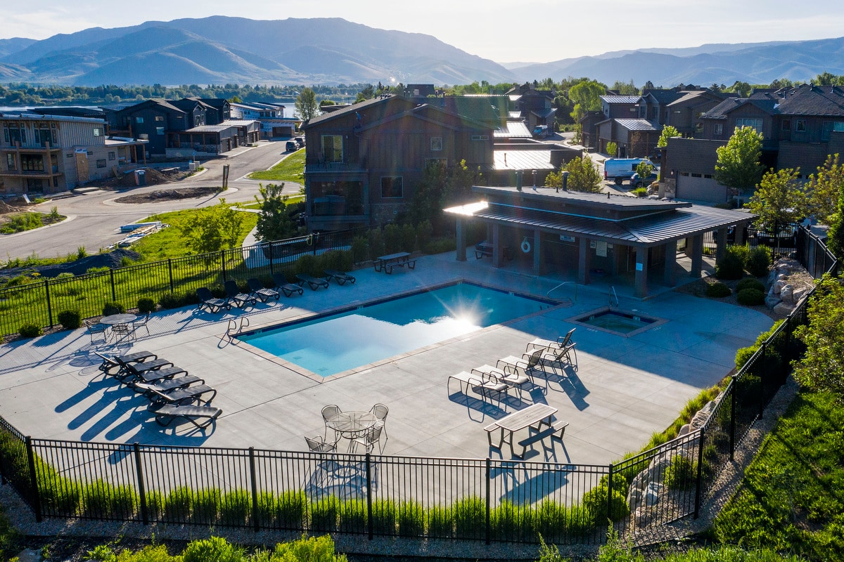 The outdoor pool area is surrounded by a stylish fence, featuring lounge chairs and shaded tables for relaxation. A hot tub is visible nearby, framed by greenery and offering views of the surrounding mountains.