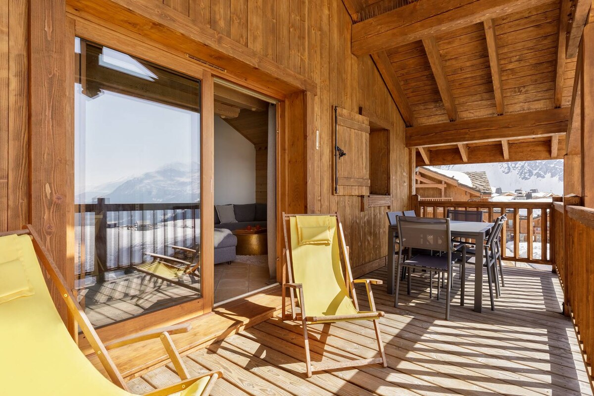 A wooden balcony is displayed, featuring yellow lounge chairs and a dining table with seating. The expansive glass doors lead to a cozy living space, showcasing a view of the surrounding mountains and valley. Overhead, the ceiling is finished with wooden beams.