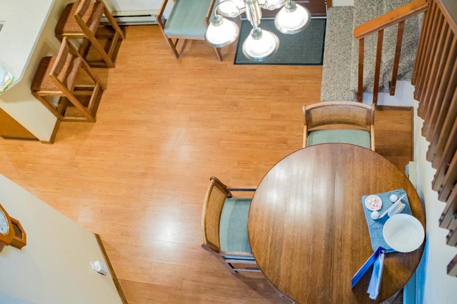 An aerial view of a cozy dining area features a round wooden table surrounded by four chairs. A chandelier with multiple lights hangs above the table, and two bar stools are positioned near the counter in the background. Warm wood flooring creates a welcoming atmosphere.