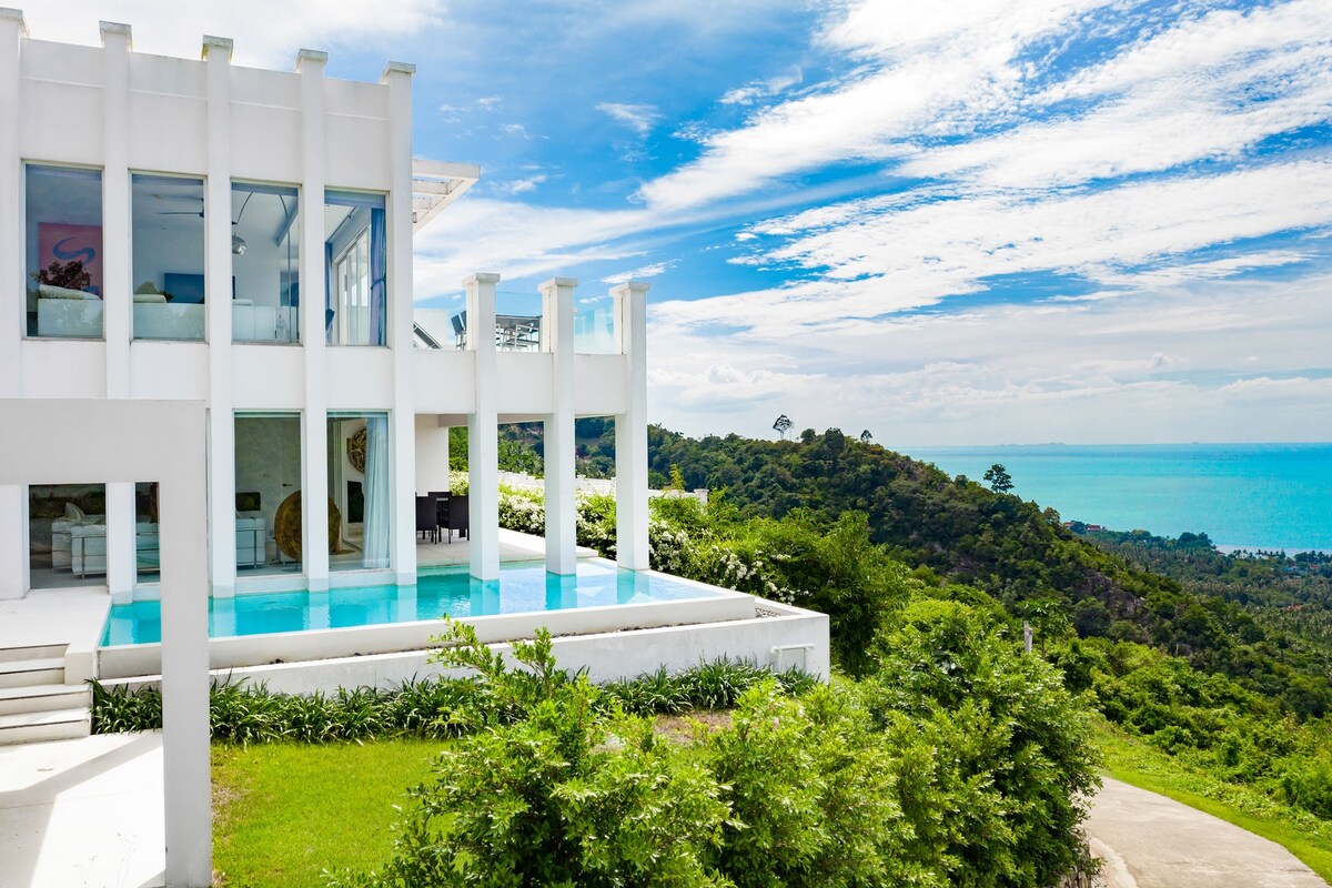 A modern property is showcased against a backdrop of rolling hills and the sea. The infinity pool reflects the sky while the expansive terrace offers views of lush greenery and the vibrant blue ocean. Sunlight enhances the clean architectural lines of the residence.
