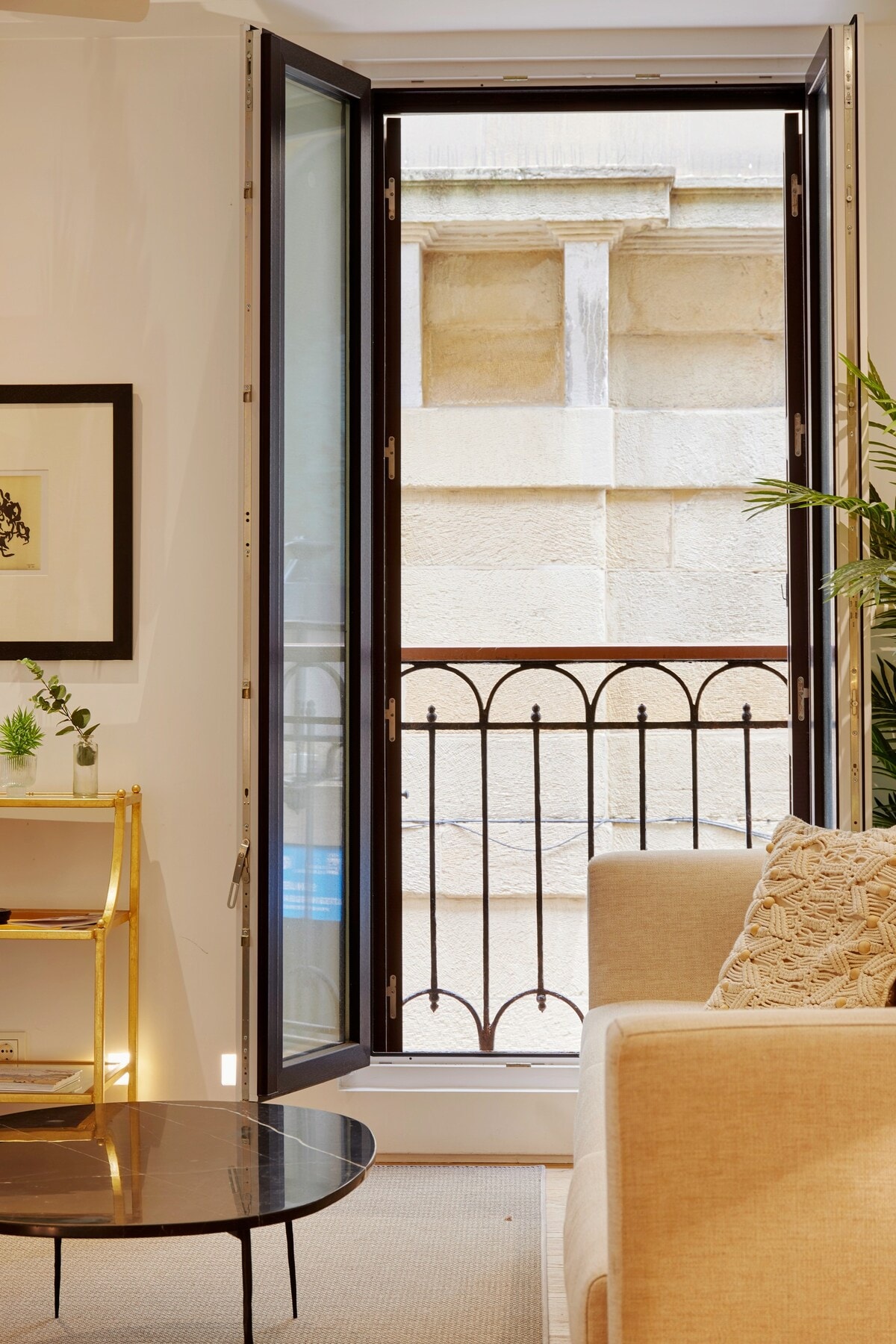 Large open windows reveal a charming balcony with ornate ironwork, allowing natural light to fill the cozy living space. A neutral-toned sofa is positioned beside a round coffee table, complementing the modern decor and creating a welcoming ambiance.