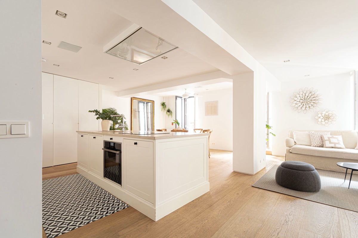 A spacious kitchen is featured with a central island, complemented by modern cabinetry and a built-in oven. Natural light fills the open-concept area, which connects to a cozy living space furnished with a light-colored sofa and a soft ottoman, creating a bright and welcoming environment.