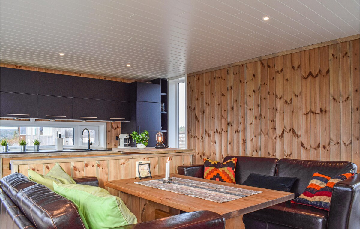 An open living area features wooden walls with a natural finish, complemented by modern cabinetry. A large wooden table is surrounded by leather sofas adorned with colorful cushions, creating a comfortable space. Large windows provide ample natural light and views of the surroundings.