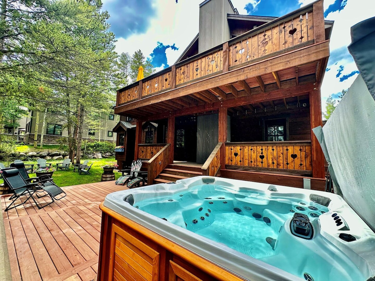 The backyard features a modern hot tub surrounded by wood decking, with lounge chairs positioned nearby. A spacious deck above offers a wooden railing and outdoor seating, with trees providing a natural backdrop. Sunlight filters through the branches, creating a serene outdoor retreat.