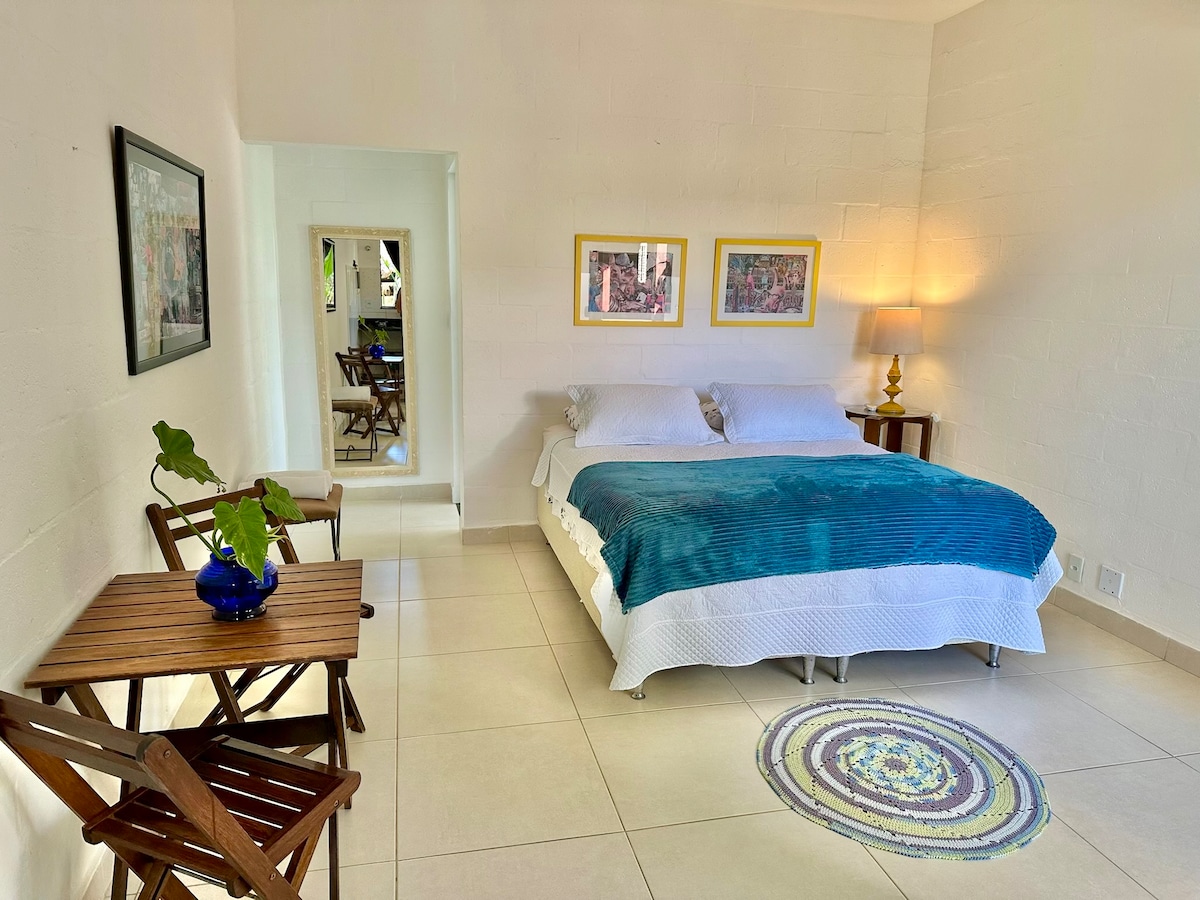 A bright and airy room features a spacious bed dressed in blue linens, complemented by two framed artworks on the walls. A small wooden table and chairs are positioned nearby, while a circular rug adds a colorful touch to the tiled floor. A mirror reflects the inviting space.