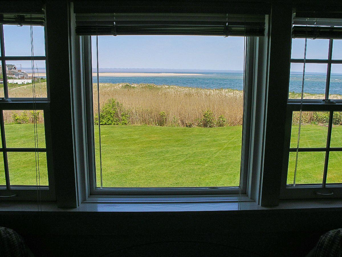 The view from the window showcases a vibrant green lawn leading to the sandy beach and the Atlantic Ocean. Sunlight brightens the scene, illuminating the gentle waves and sandy shores in the distance, framed by the window's white casing and thin blinds.