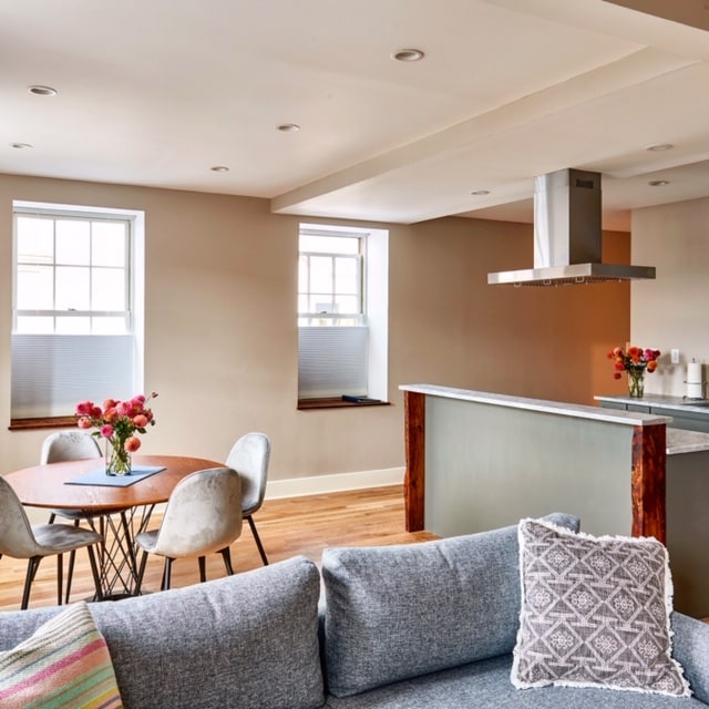A bright living area features a textured sofa and a circular dining table surrounded by four chairs. Natural light filters through two large windows, and a modern kitchen area with a sleek countertop is visible in the background. Fresh flowers add a touch of color.