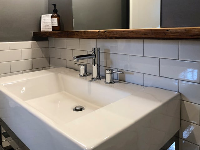 A modern sink area with a spacious white basin is showcased against a backdrop of white subway tiles. A wooden shelf above the sink displays a bottle of amenities. The sleek silver fixtures contrast with the darker wall, creating an elegant and functional space.