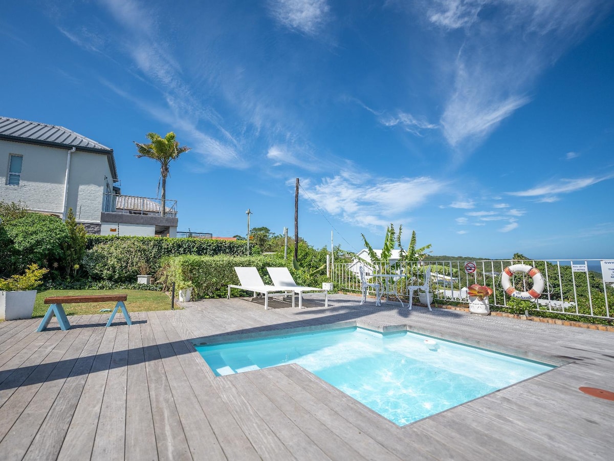 A private pool is framed by a wooden deck, surrounded by lush greenery and a sunny sky. Two white lounge chairs are positioned beside the pool, while a small table adds a touch of functionality to the area. Scenic views can be seen in the background.