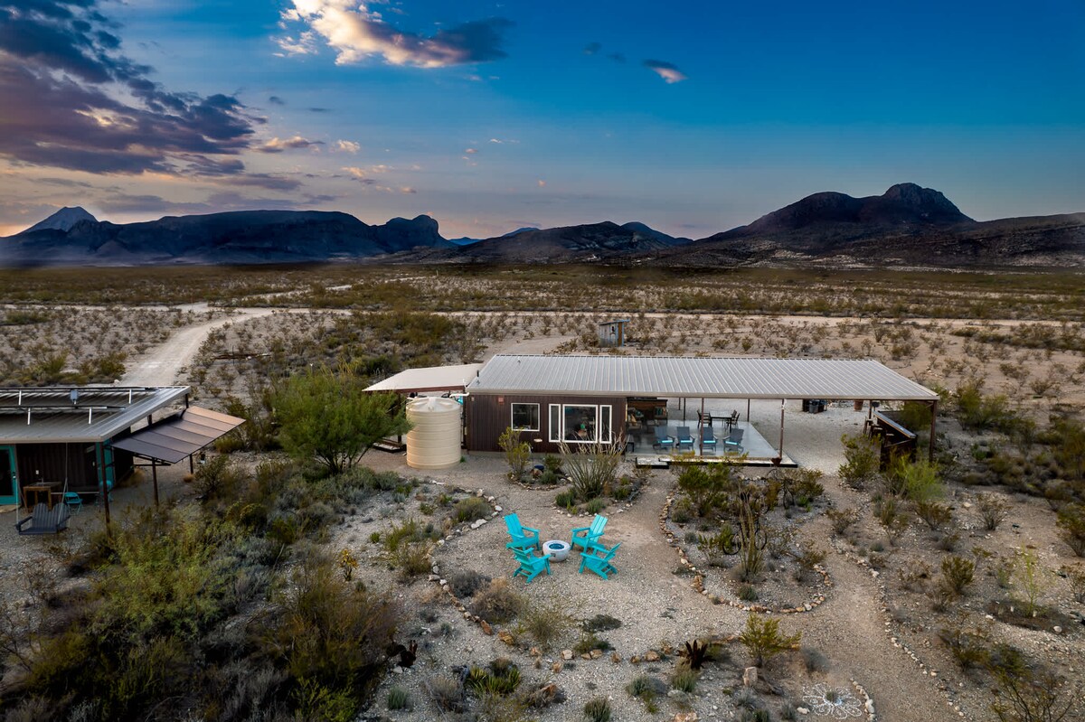 The off-grid rustic modern home is nestled within the desert landscape, surrounded by mountain views. The property features a covered porch, outdoor seating, and a designated fire pit area, all set against the backdrop of expansive skies. Solar panels are visible on the roof.