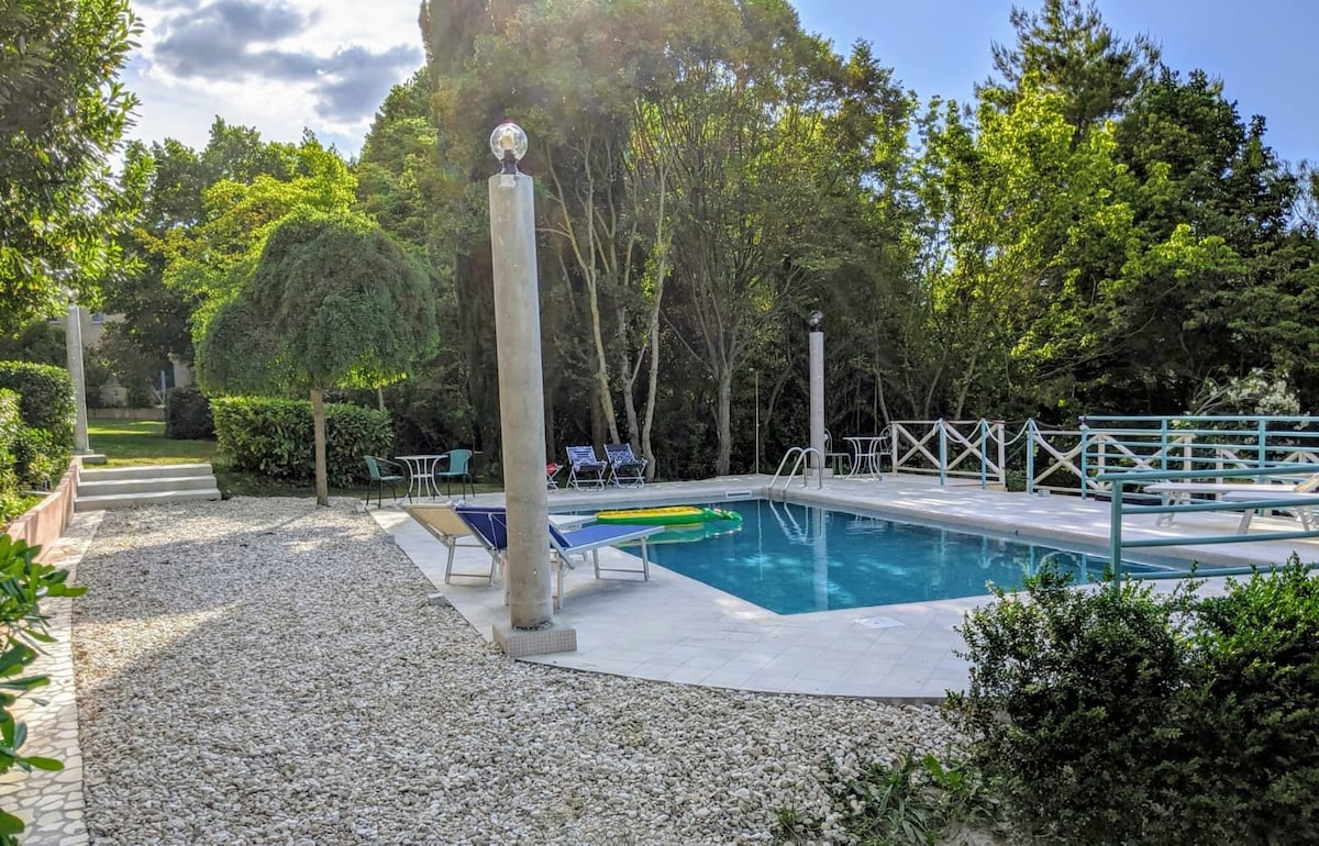 A serene outdoor pool area is surrounded by lush greenery. The pool is flanked by lounge chairs and equipped with safety features. A stone pathway leads to the space, adorned with well-manicured plants creating a relaxing atmosphere.