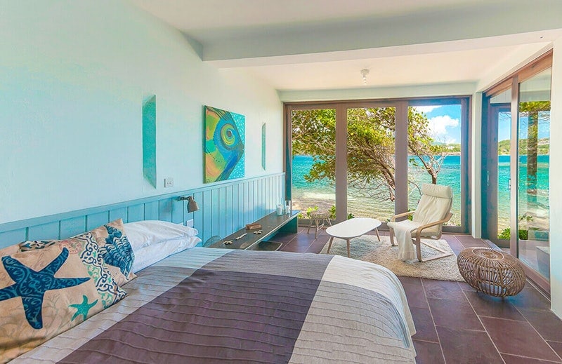 A spacious bedroom features a comfortable bed with decorative pillows, complemented by modern art on the walls. Large glass doors provide views of the ocean and allow natural light to fill the room. A cozy seating area with a coffee table is visible nearby.