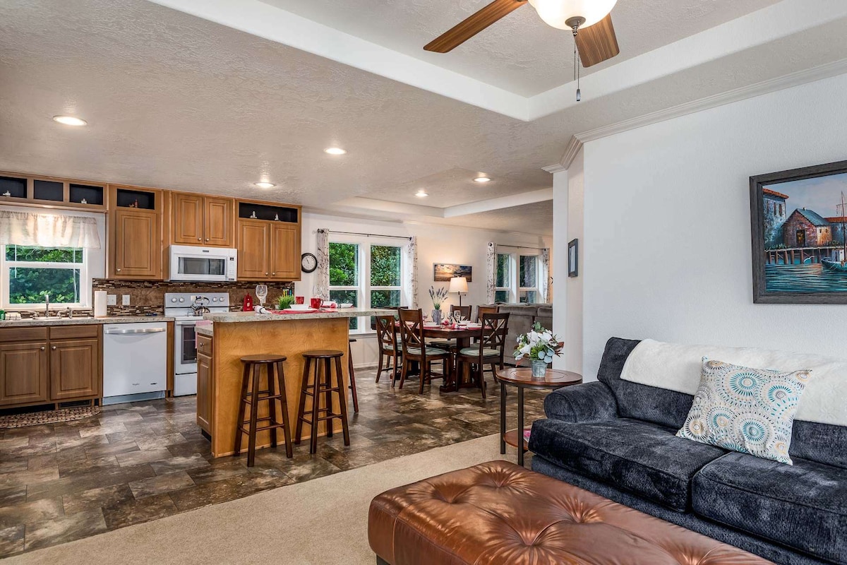 An open-concept kitchen and living area features modern appliances and a central island with seating. Natural light filters in through large windows, illuminating the space. Comfortable seating is arranged around a table set for dining, while artwork and decor add a touch of charm.