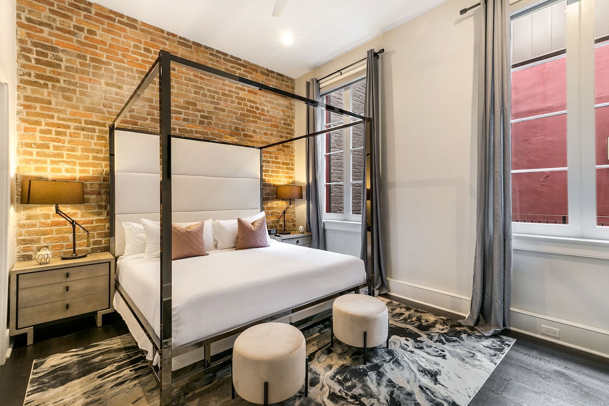 A spacious master bedroom features exposed brick walls and original wooden beams. A king-size bed, dressed in luxury linens, is centered in the room, complemented by two cushioned stools. Natural light enters through large windows, adorned with sheer curtains.