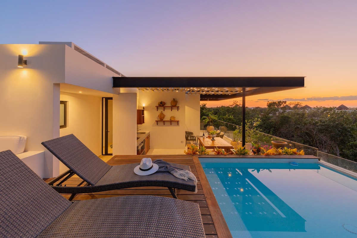 Two lounge chairs are situated beside a rooftop infinity pool, showcasing an open-air dining area and a well-equipped kitchen. A sunset casts a warm glow over the tranquil jungle landscape, enhancing the serene atmosphere of this outdoor space.