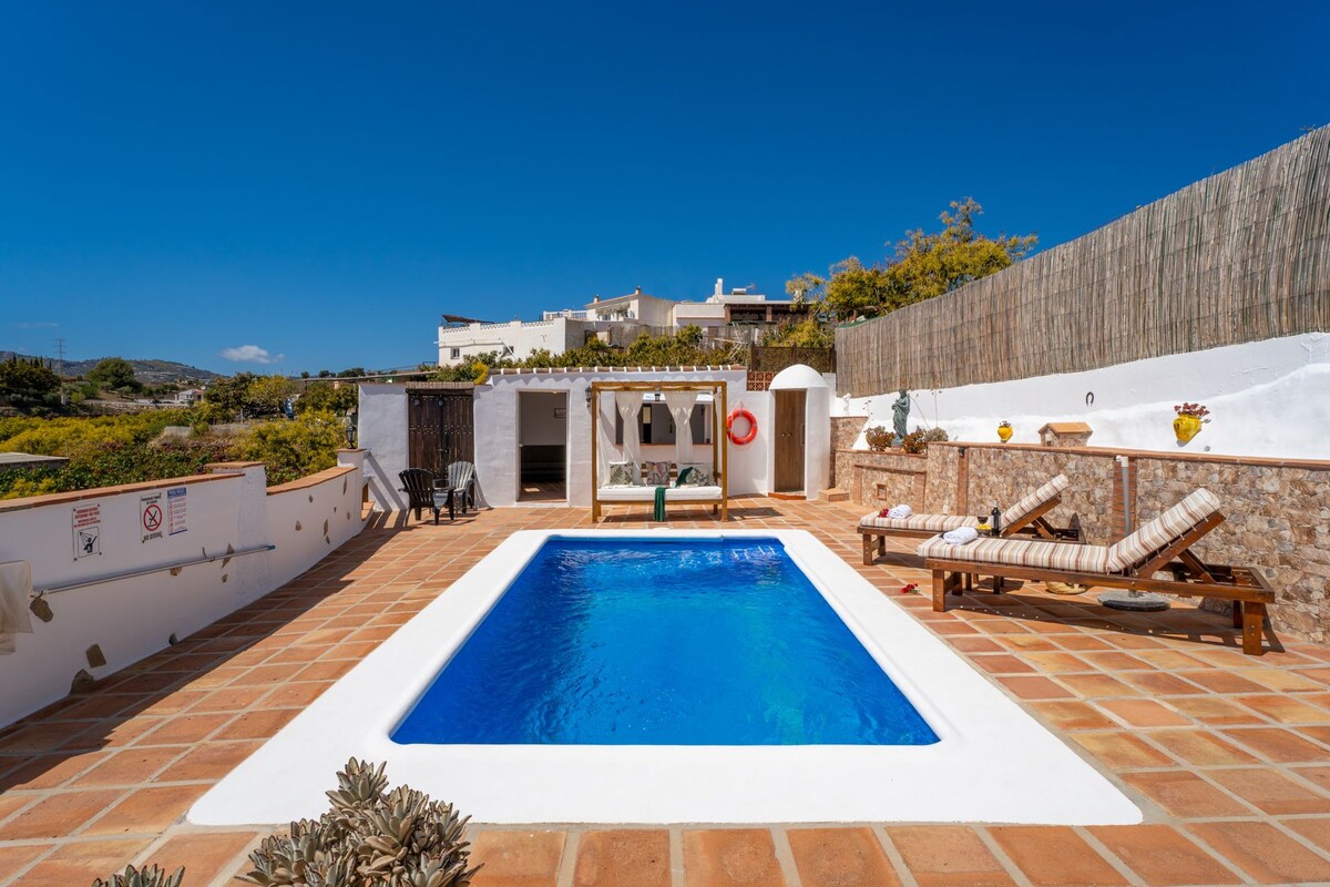 A private swimming pool is framed by tiled terraces and complemented by sun loungers. The surrounding walls display natural stone features, and a wooden privacy screen provides shade. Lush greenery and potted plants enhance the outdoor space, under a clear blue sky.