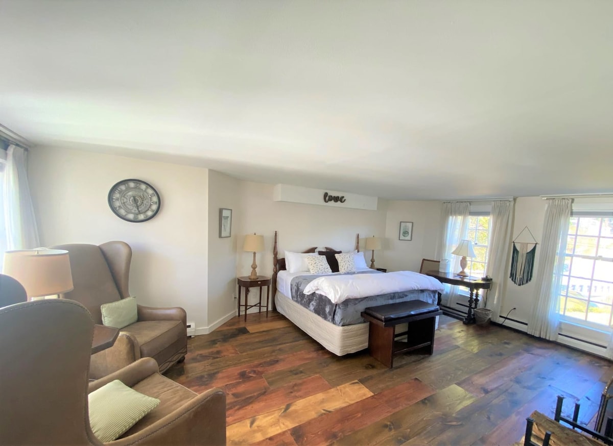 The spacious room features a king bed with crisp linens, complemented by two bedside lamps. A cozy sitting area includes a wingback chair and a small table. Natural light floods the space through large windows, and warm wood flooring enhances the inviting atmosphere.