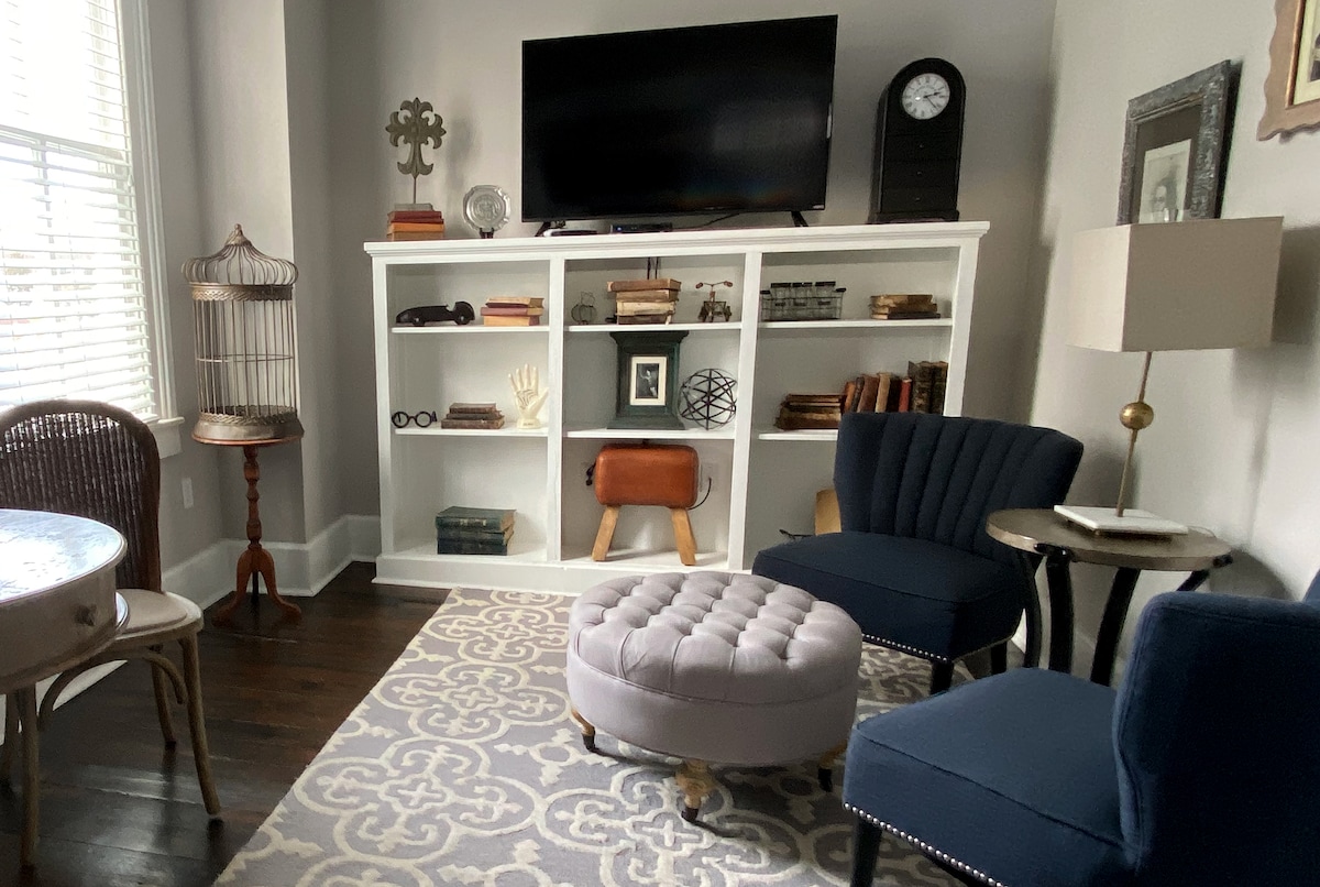 An inviting sitting area features two blue upholstered chairs and a tufted ottoman, set on a patterned rug. A white built-in bookshelf displays curiosities and books, while a flat-screen TV is mounted above. Natural light enters through a window with adjustable shades.