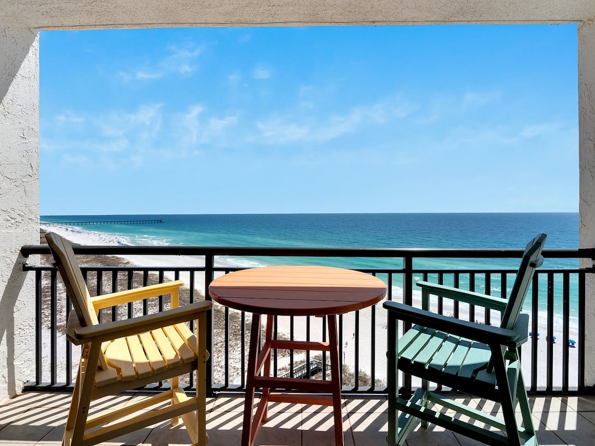 A private balcony showcases two colorful Adirondack chairs alongside a round wooden table, overlooking expansive views of the Gulf. The bright blue sky complements the sparkling water, and the sandy beach can be seen in the distance, enhancing the tranquil seaside setting.