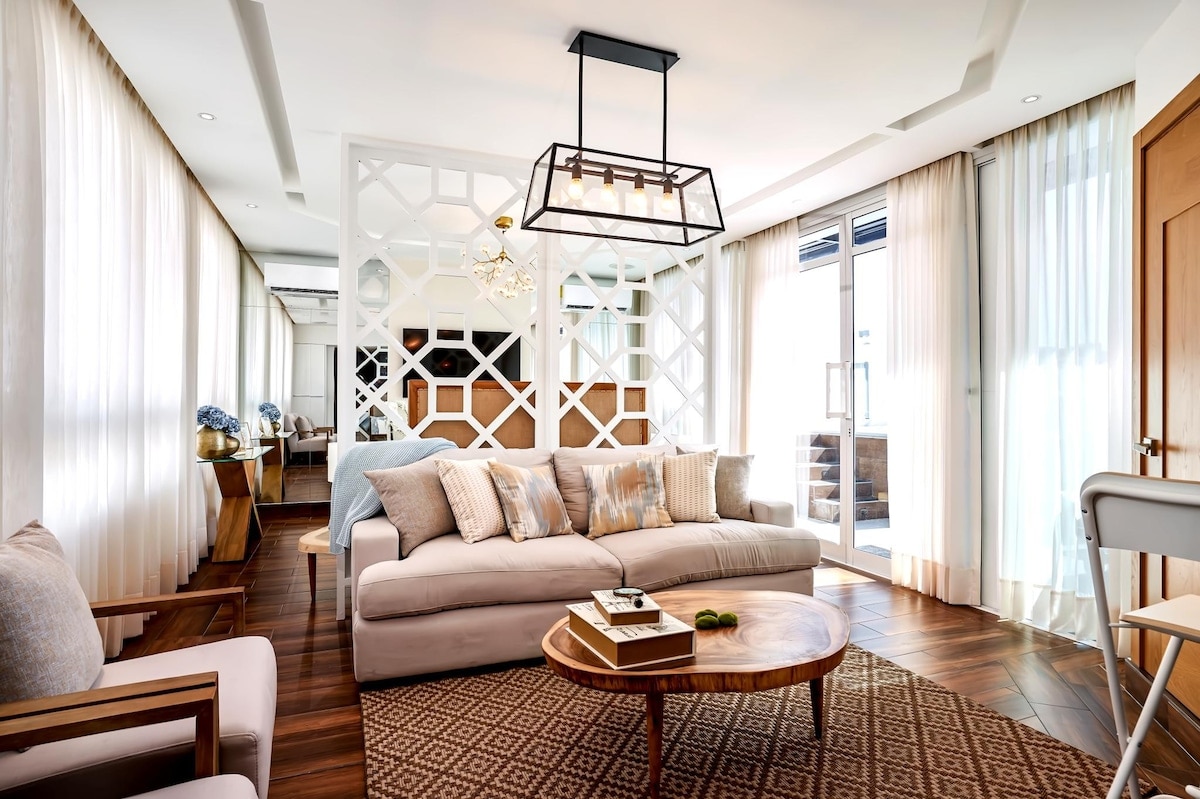 A modern living area features a light, neutral color palette with a spacious sofa adorned with cushions. A round wooden coffee table sits in front, accompanied by a statement light fixture above. Large windows draped in sheer curtains allow natural light to brighten the room.