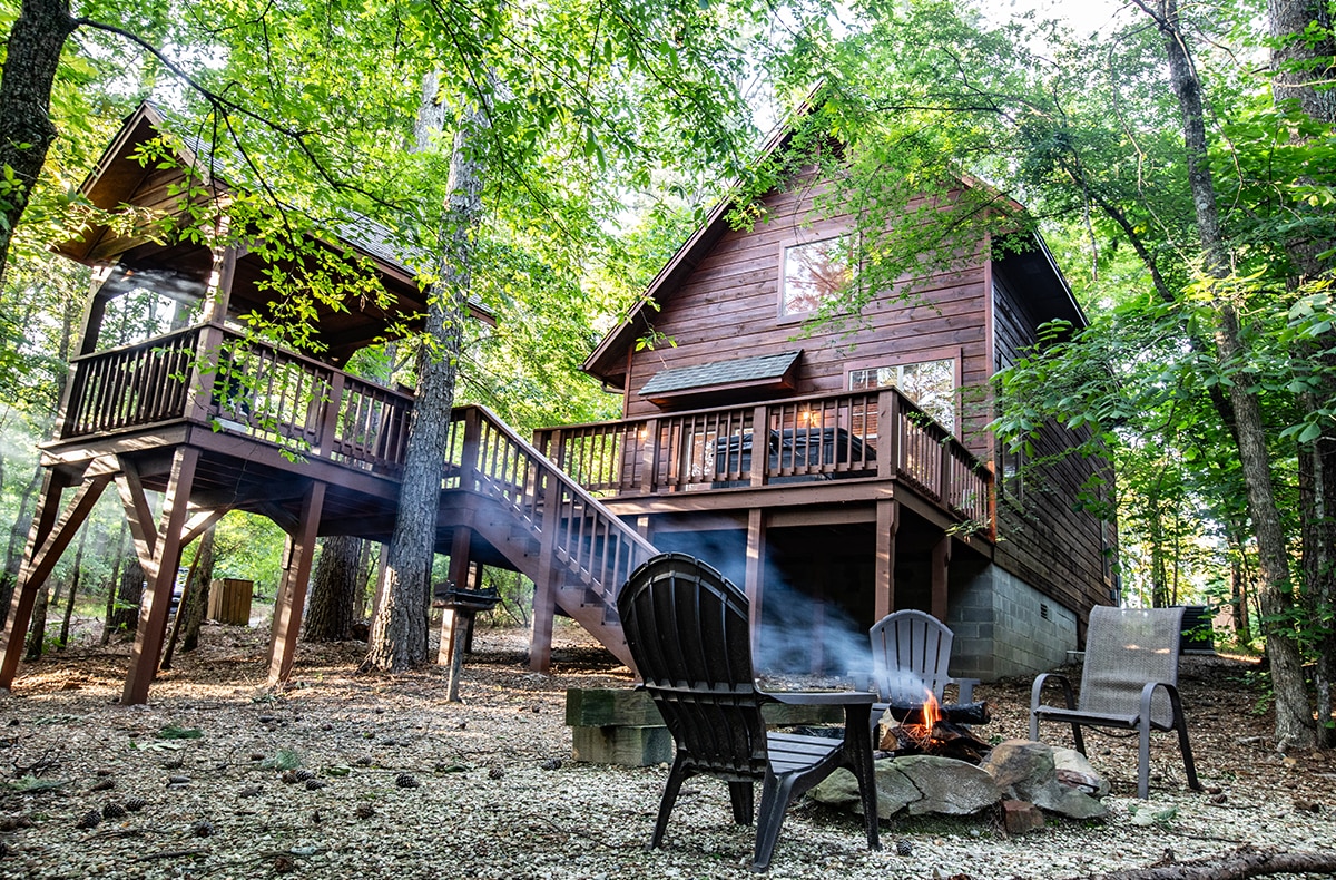A secluded cabin nestled among lush greenery features a wooden exterior and is surrounded by tall trees. An outdoor fire pit with seating is positioned in front, providing a cozy space for gatherings. A staircase leads to an upper deck, enhancing outdoor relaxation.