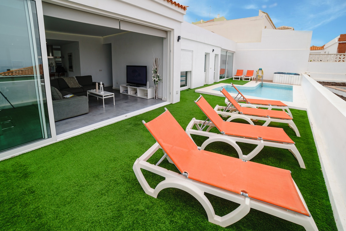 An outdoor area features multiple loungers arranged around a private pool, with bright orange cushions offering a pop of color against the green grass. Sliding glass doors lead into the living space, maintaining a seamless connection between indoor and outdoor relaxation.