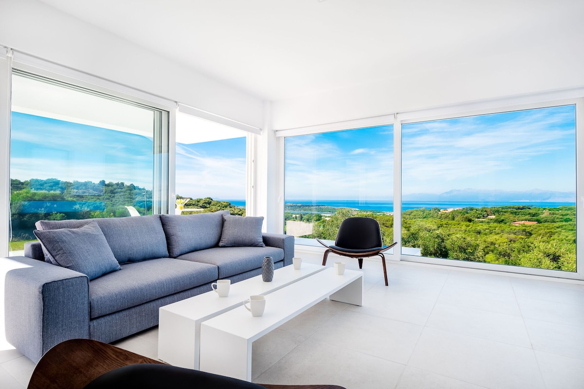 A spacious lounge area features a large gray sofa and a minimalist white coffee table. Expansive glass windows offer panoramic views of lush greenery and the coastline, allowing natural light to fill the room.