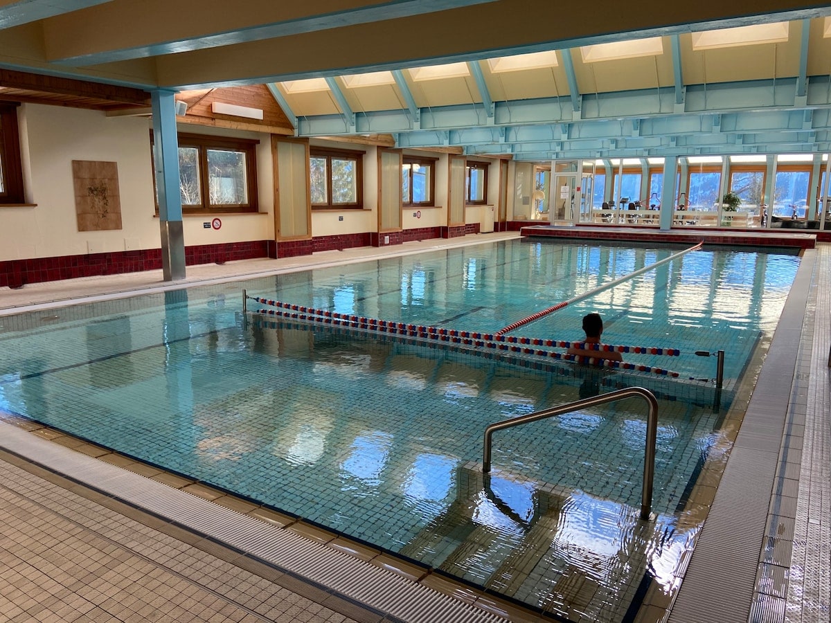 An indoor swimming pool is illuminated by natural light from large windows, revealing views of the surrounding landscape. The pool area features tiled flooring, a railing for easier access, and marked lanes in the water. Comfortable seating is arranged around the space.