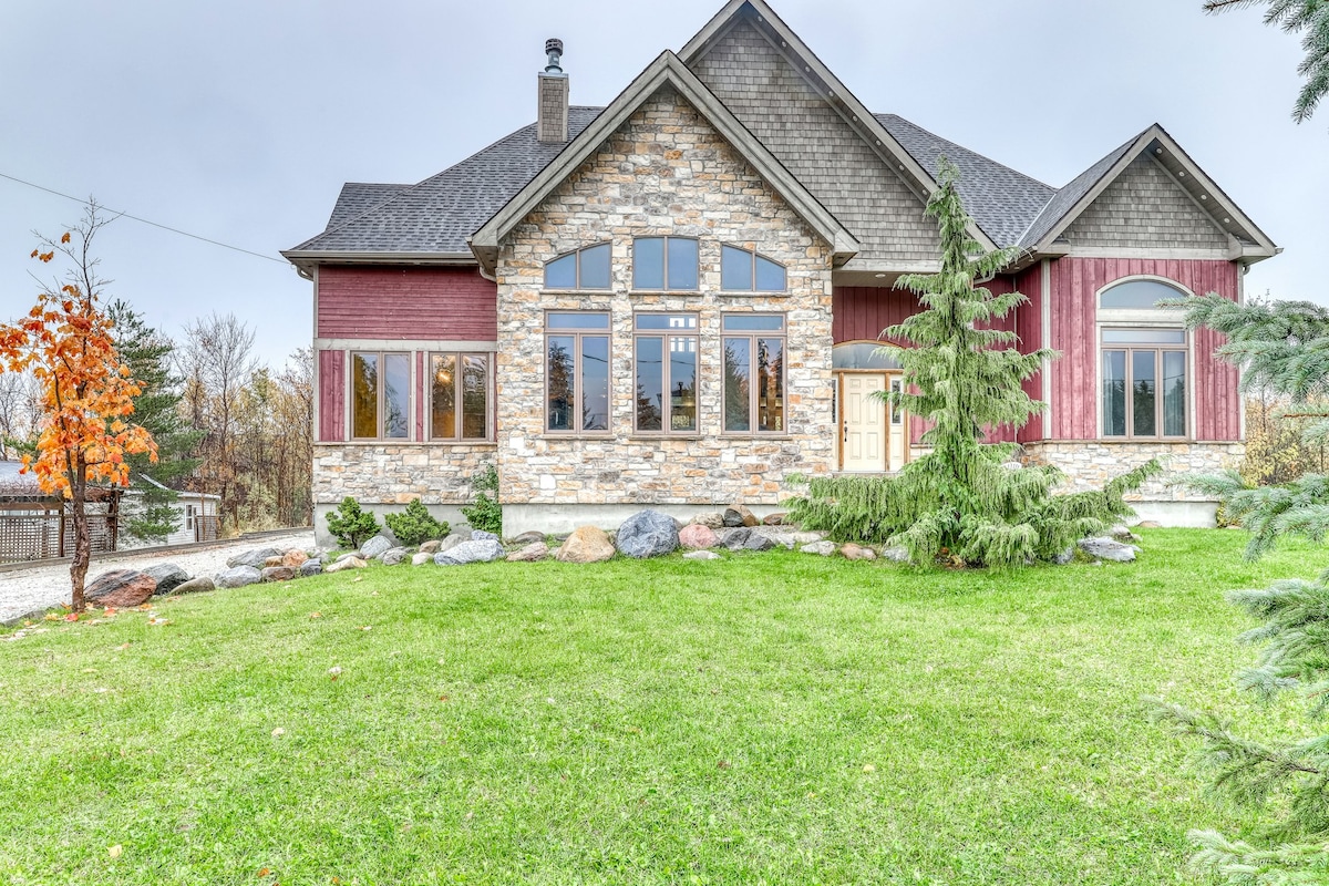 The exterior of the custom-built ski chalet is showcased, featuring a mix of stone and wood finishes. Large windows provide ample light and a view of the surrounding landscape, while a neatly maintained lawn and strategic landscaping elements enhance the home's appeal.