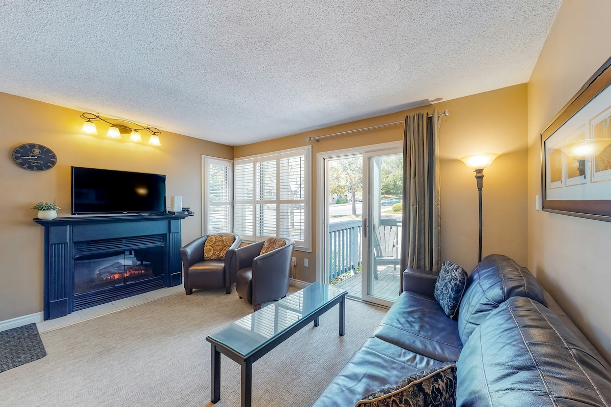 A living area features a comfortable sectional leather sofa and two armchairs, arranged around a glass coffee table. A television is mounted above a fireplace, and sliding glass doors lead to a private deck, allowing natural light to fill the space.