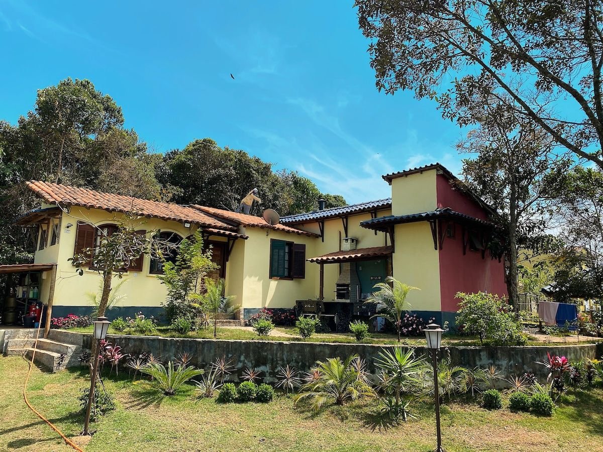 A charming two-part house is set against a backdrop of greenery, featuring a tiled roof and vibrant exterior walls. The well-maintained garden is enhanced by a variety of plants and shrubs, and pathway lights are visible along the lawn.