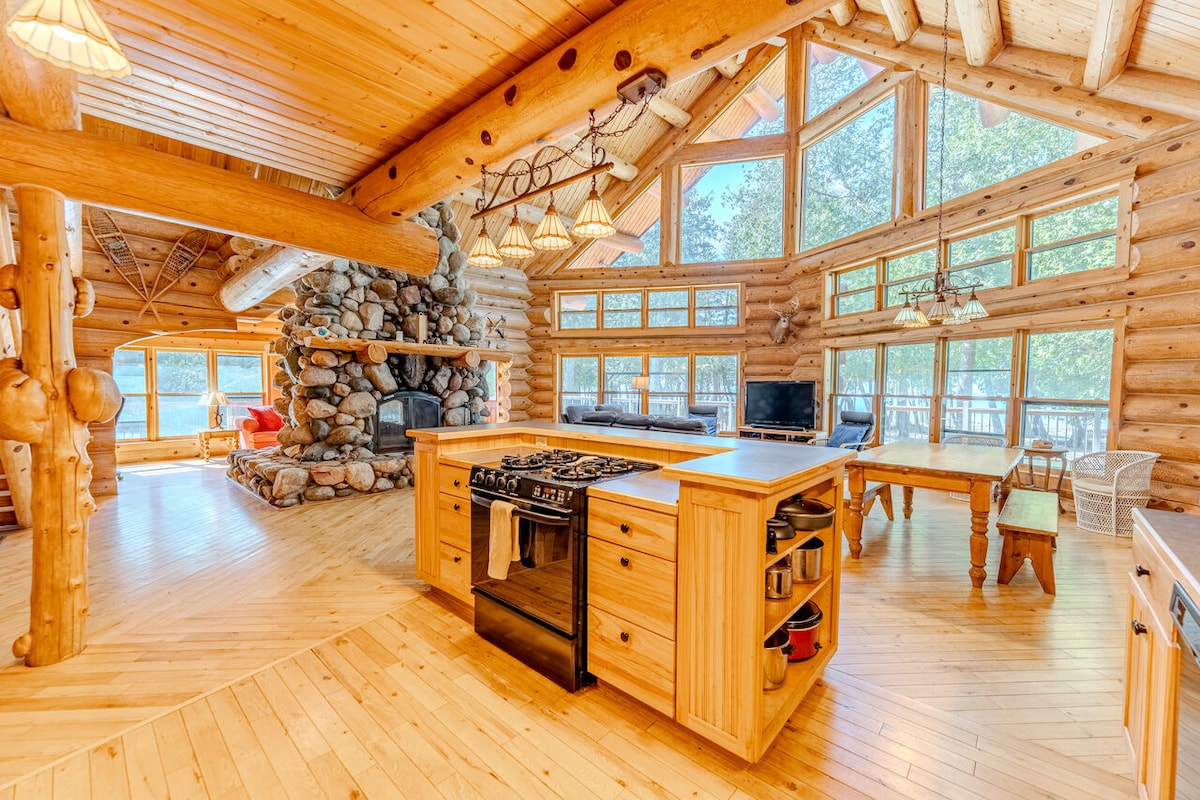 The open-concept kitchen features a central island with bar seating, surrounded by wooden cabinetry. Large windows allow natural light to flood the space, highlighting the intricate log construction and a stone fireplace in the background. A dining area is visible, offering a welcoming atmosphere.