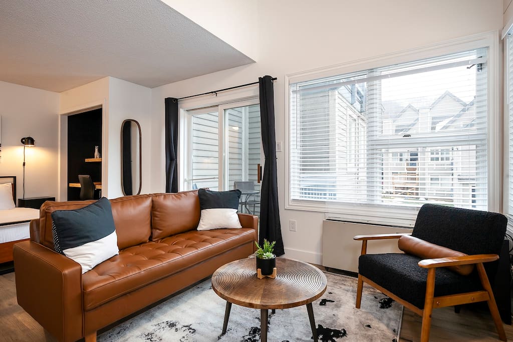 The living area features a brown leather sofa and a black armchair positioned around a round wooden table. Natural light enters through large windows framed by black curtains, showcasing views of the outdoors. A neutral-colored area rug anchors the space.